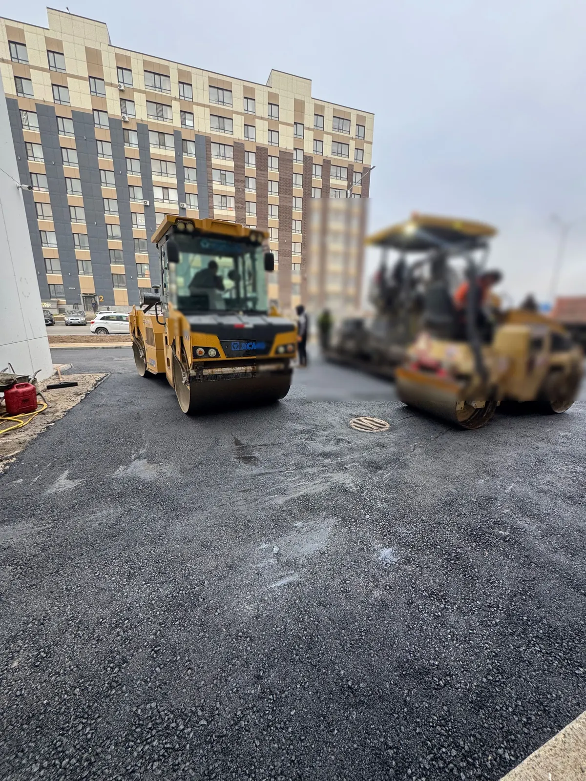 Два дорожных катка уплотняют свежий асфальт на площадке у жилого дома в Астане